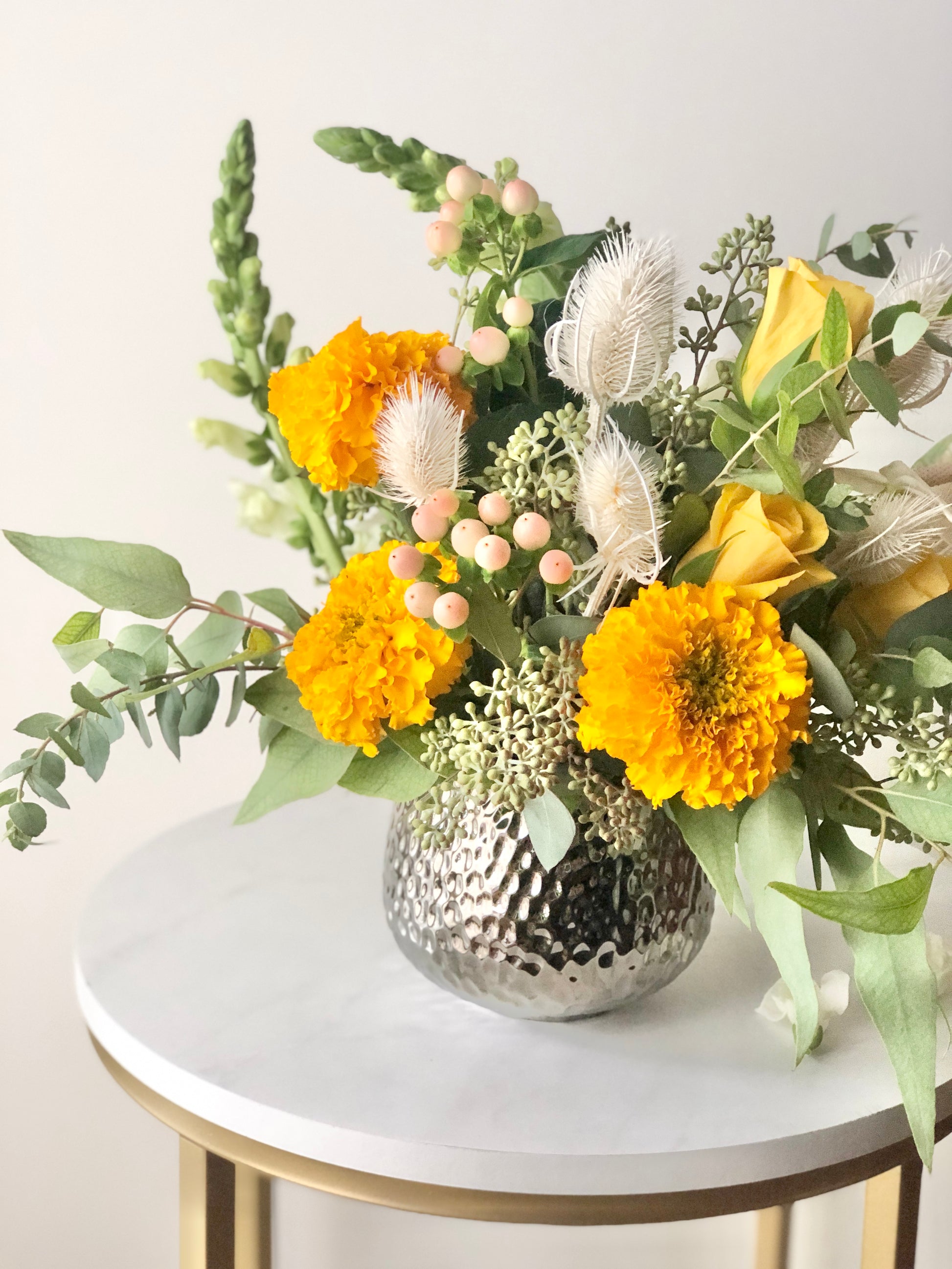 View of Marigolds in Bohemian style floral arrangement with Soft Orange, yellow, cream flowers with loose sage foliage in a luxury vase