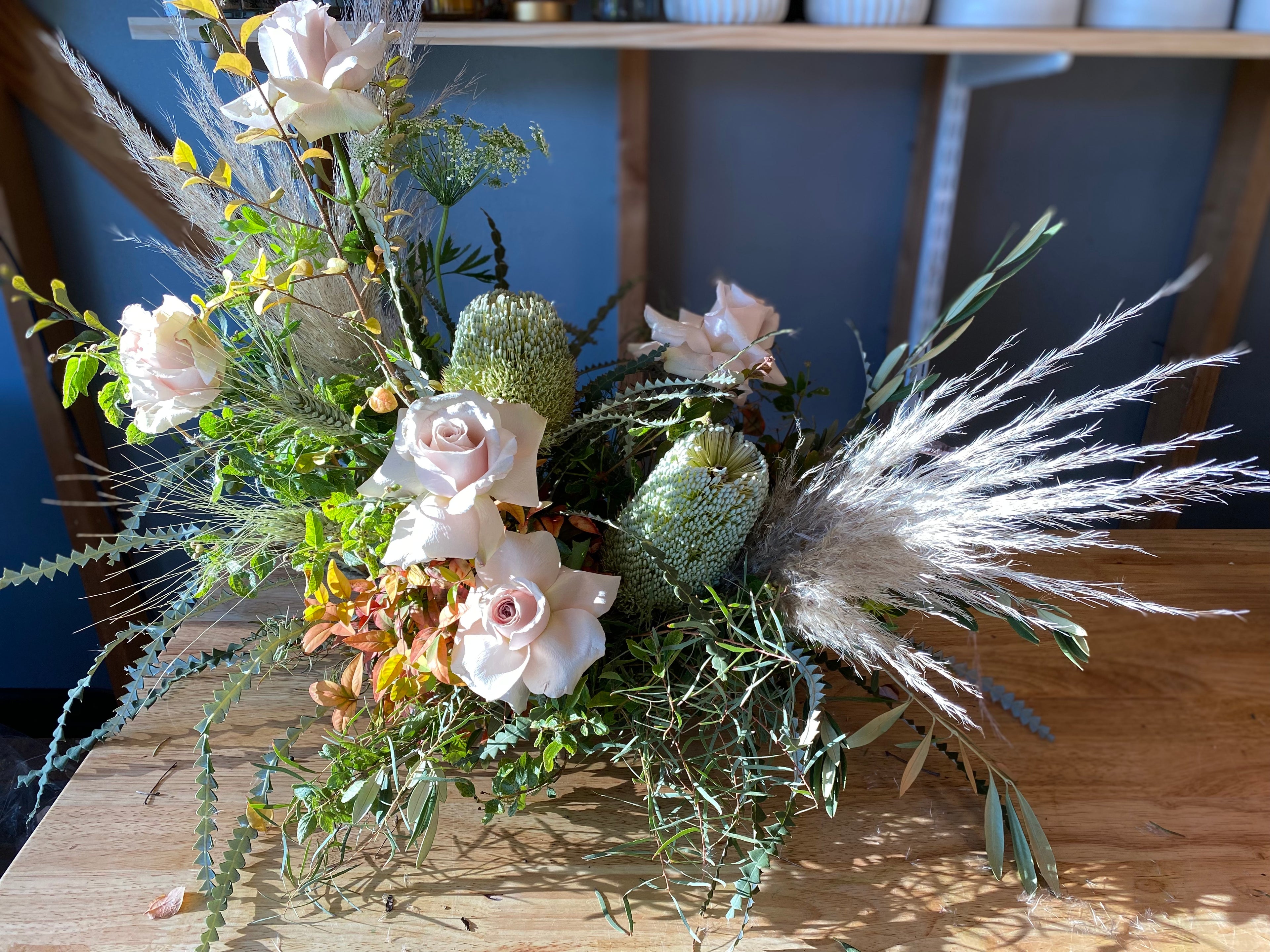 Modern Floral Design of sage green banksia and blush rose with pampas grass and olive greenery on a wooden workbench